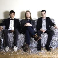 Three caucasian men sitting on a grey couch with shredded material decorating it