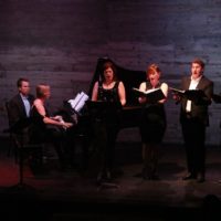 A photo of 4 singers, a pianist, a page turner and a violinist performing in a dark space