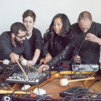 Four musicians crowd around electronics on a table, messing with knobs and wires