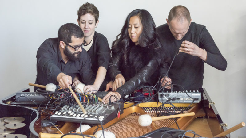 Four musicians crowd around electronics on a table, messing with knobs and wires