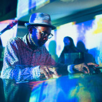 Noel Brass Jr adjusts a DJ Deck as he is overcast with colorful projections