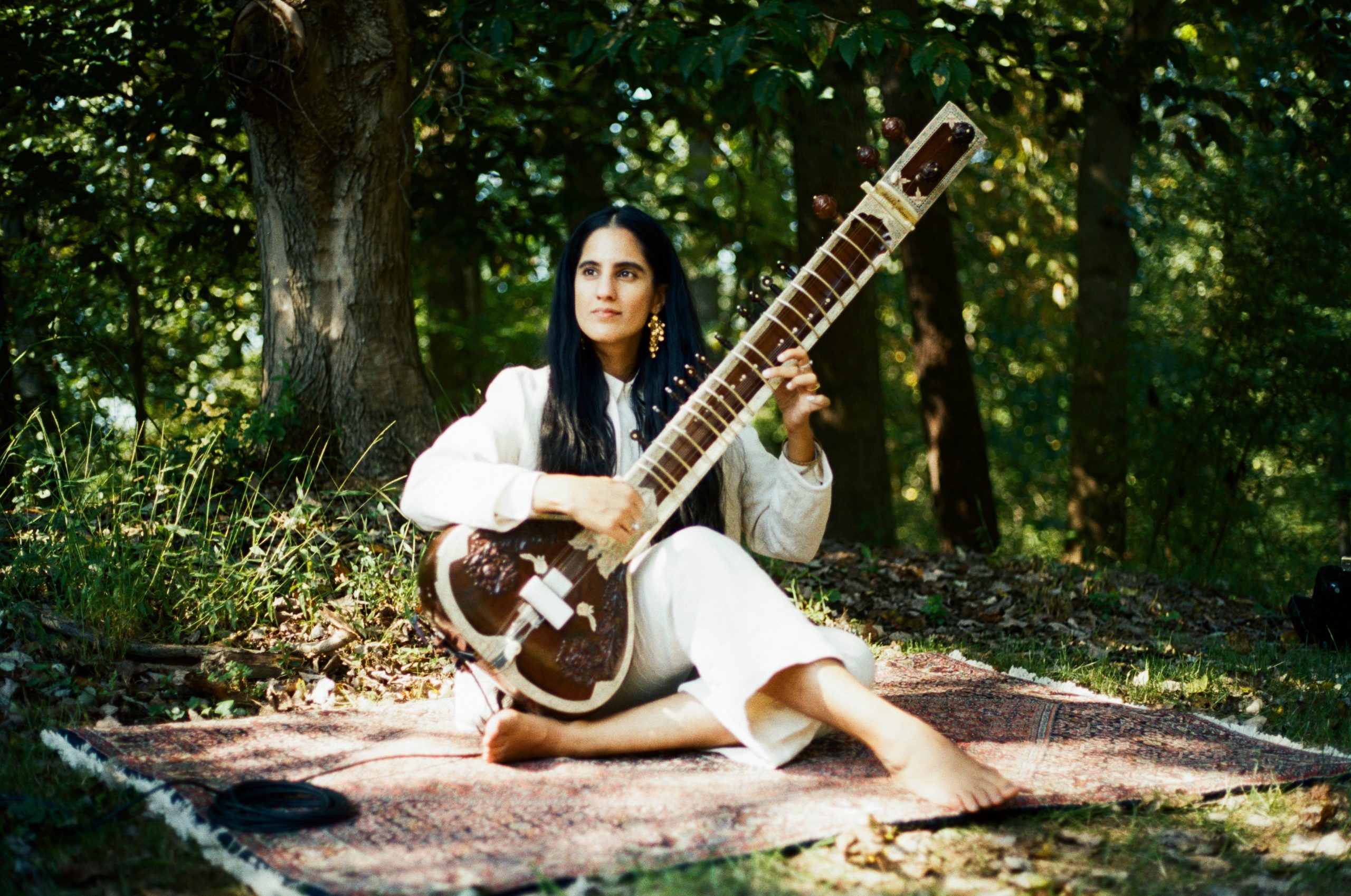 Ami Dang outside playing a sitar in front of several trees.