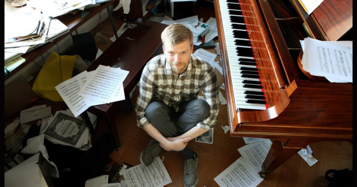 Andrew Norman sitting by his piano with pages of scores scattered on the floor.